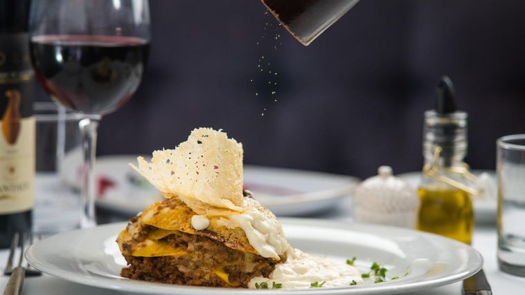 Lasagne auf einem Teller, garniert mit Käse und Kräutern. Im Hintergrund ein Glas Rotwein und eine Flasche Olivenöl. Gewürze werden darüber gestreut.