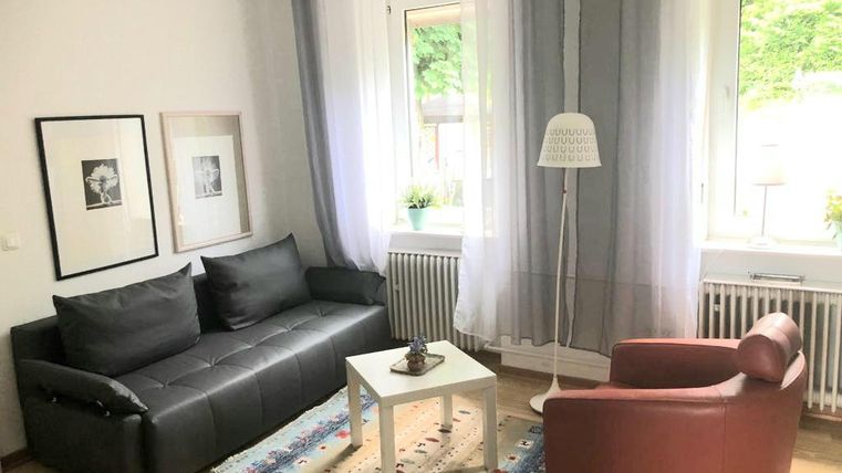 Ein gemütliches Wohnzimmer mit einer schwarzen Couch, einem roten Sessel und einem kleinen Tisch. Große Fenster lassen viel natürliches Licht herein.