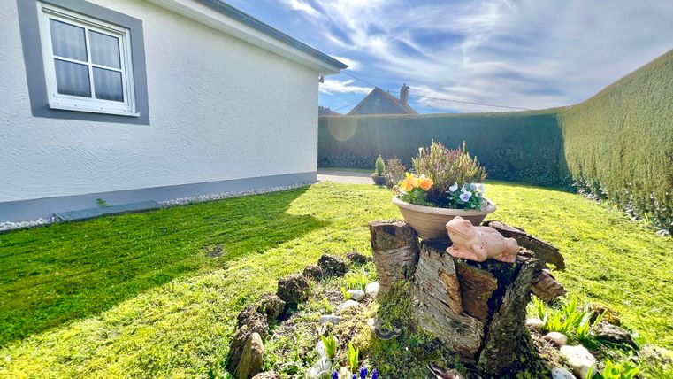 A well-maintained garden with a tree stump decorated with flowers. In the background, there is a bright house and a blue sky.
