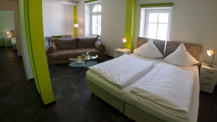 Ein gemütliches Zimmer mit einem Doppelbett und einem Sofa. Die Wände sind in frischem Grün gestrichen, und es gibt große Fenster, die viel Licht hereinlassen.