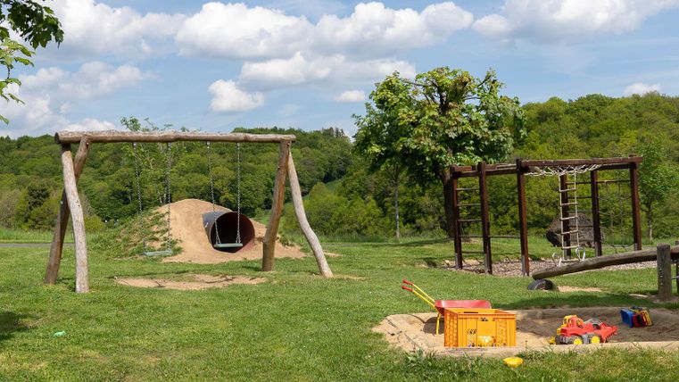 Een speelplaats met een wip, een glijbaan en een zandvlak. Op de achtergrond zijn groene bomen en een blauwe lucht te zien.