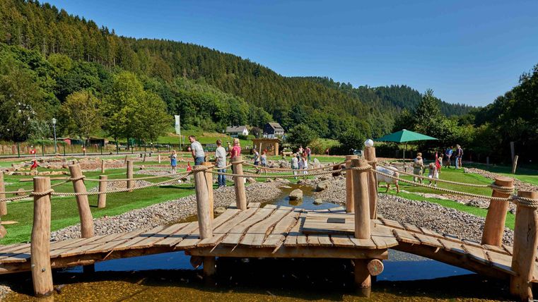A picturesque wooden cart crosses a small waterway. In the background, green hills and visitors can be seen enjoying nature.