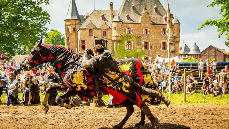 Een ridder in volle wapenrusting rijdt op een prachtig paard met een kleurrijke mantels. Op de achtergrond zijn een historische burcht en een grote menigte te zien.