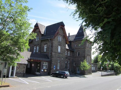 A large three-story building with two tower-like projections, gable roofs, and white windows. The main structure is essentially made of red rubble stone and has a separate outbuilding. In front of the building, a dark car is parked.