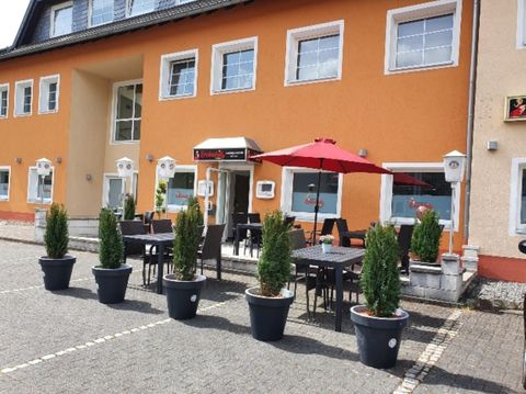 Een modern gebouw met oranje gevel en grote ramen. Buiten staan tafels met stoelen en een rode parasol, omringd door planten in zwart-witte potten.