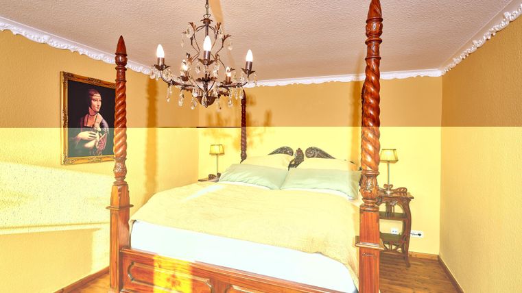 A cozy bedroom with a four-poster bed and an elegant chandelier. The walls are painted in warm yellow and there is a classic painting on the wall.