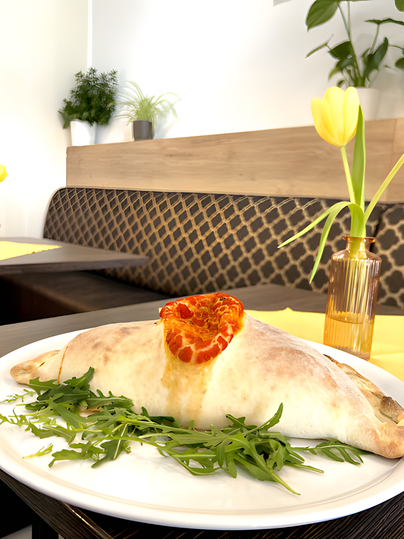 Ein köstlicher Calzone auf einem weißen Teller, garniert mit Rucola und einer Scheibe Tomate. Der Hintergrund zeigt eine gemütliche Sitzecke mit Pflanzen und frischen Tulpen.