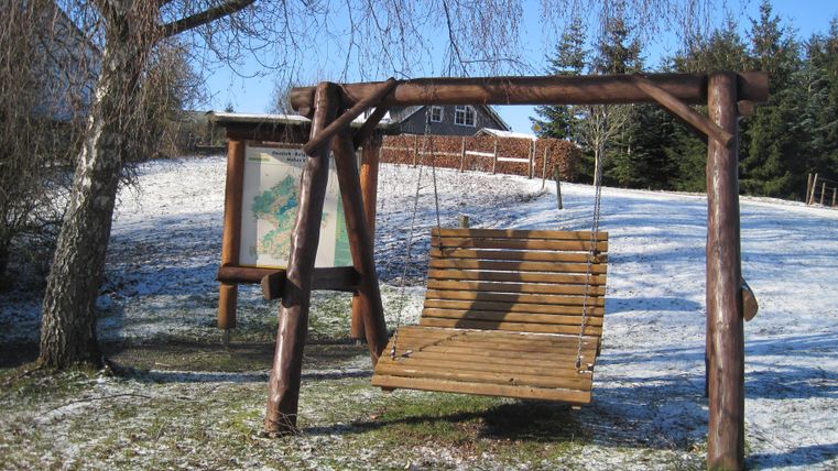 Holzschaukel und Infotafel in einem verschneiten Park. Im Hintergrund sind Bäume und ein Haus zu sehen. Der Himmel ist klar und blau.