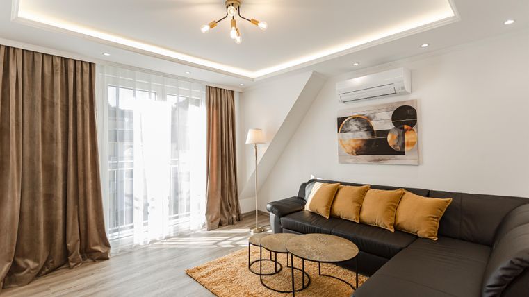 Ein modernes Wohnzimmer mit einer bequemen schwarzen Couch und dekorativen Kissen. Große Fenster lassen viel Licht herein und sorgen für eine helle Atmosphäre.