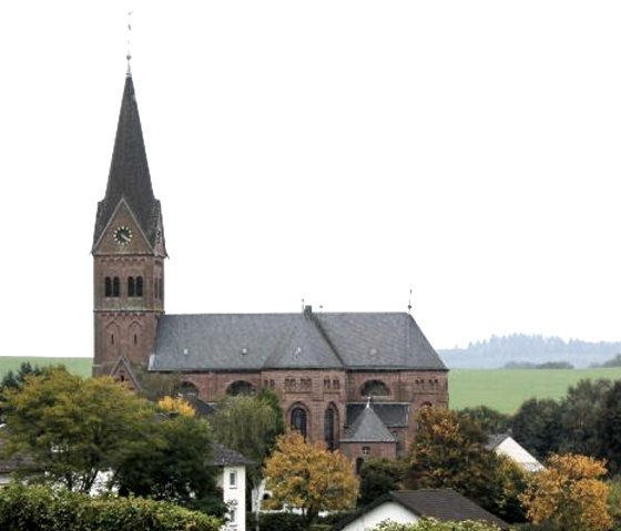 Kirche mit hohem Turm in l&auml;ndlicher Umgebung, umgeben von B&auml;umen und H&auml;usern. Der Himmel ist bew&ouml;lkt., &copy; Touristik GmbH Gerolsteiner Land