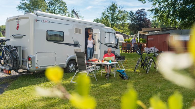 A motorhome is parked in a green campsite. People are sitting at an outdoor table and enjoying their time.