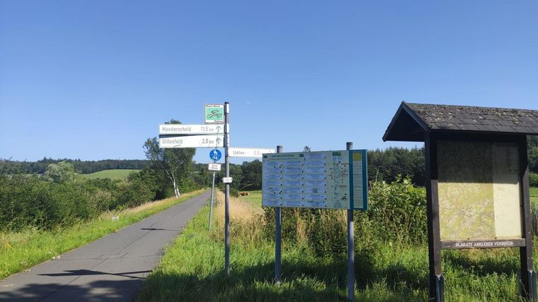 Ein ruhiger Weg mit grünen Wiesen und einem klaren blauen Himmel. Auf der Seite stehen Wegweiser und Informationstafeln.