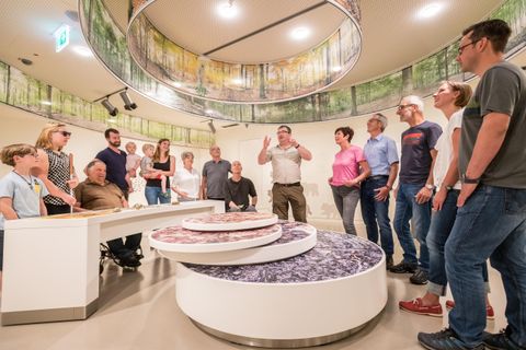 Eine Gruppe von Menschen hört einem Vortrag in einem modernen Ausstellungsraum zu. Im Mittelpunkt steht ein runder Tisch mit einer interaktiven Präsentation.