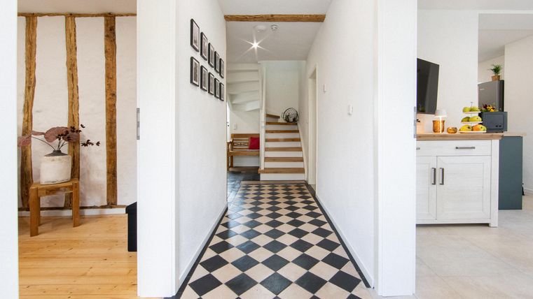 Ein heller Flur mit einem Schachbrettmuster auf dem Boden. Im Hintergrund sind Treppen und ein modernes Wohnzimmer sichtbar.