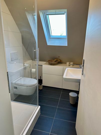 Een moderne badkamer met een douche en een raam. De muren zijn in lichte kleuren gehouden en de vloer is voorzien van donkere tegels.