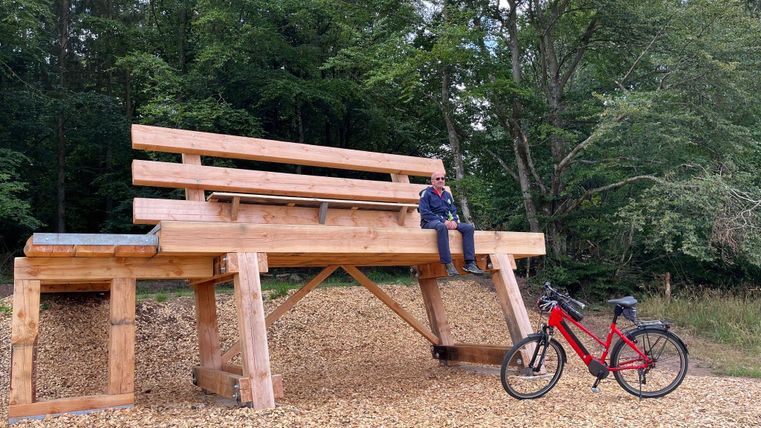 A oversized wooden bench stands in the forest, next to it a red bicycle. The bench is placed on wood shavings.