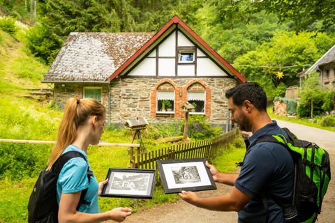 Zwei Personen betrachten Bilder vor einem alten Haus inmitten der Natur. Das Gebäude hat einen rustikalen Charme mit einem Garten und viel grünem Umfeld.