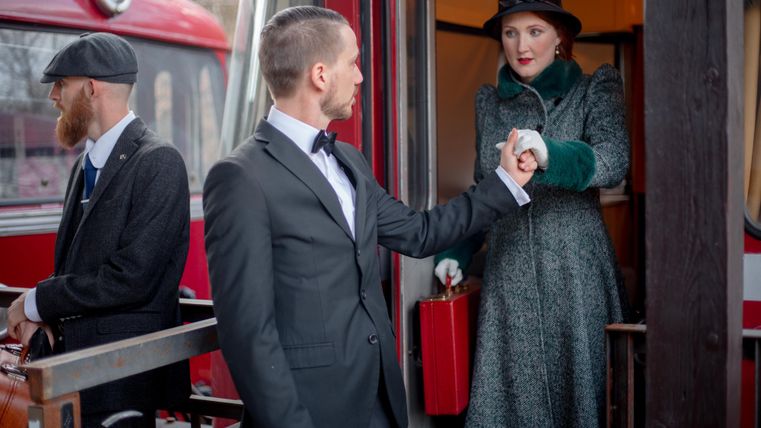 Ein Mann in einem Anzug reicht einer Frau in einem Wintermantel die Hand, während sie an einem roten Zug stehen. Eine weitere Person in einem Hut und Anzug steht daneben.