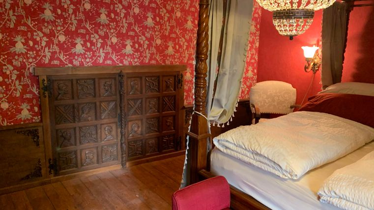 A cozy bedroom with a large four-poster bed and red wallpaper. In the background, there is a wooden wardrobe and an elegant armchair.