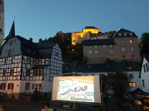 Een schilderachtige plek met historische gebouwen en een groot scherm voor een openluchtbioscoop. Op de achtergrond straalt een kasteel in de schemering.