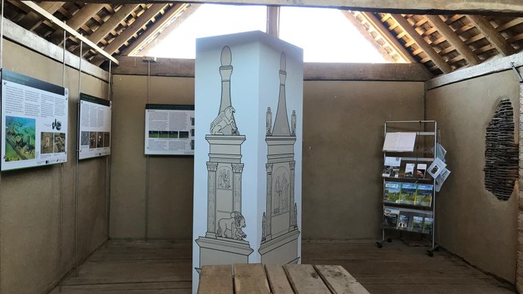 Innenansicht eines Ausstellungsraums mit Holzboden, Infotafeln an den Wänden und einem zentralen Display mit Monumentzeichnungen.