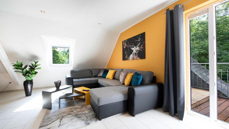 Ein modernes Wohnzimmer mit einem grauen Sofa und bunten Kissen. Große Fenster bieten einen Blick ins Freie und lassen viel Licht herein.