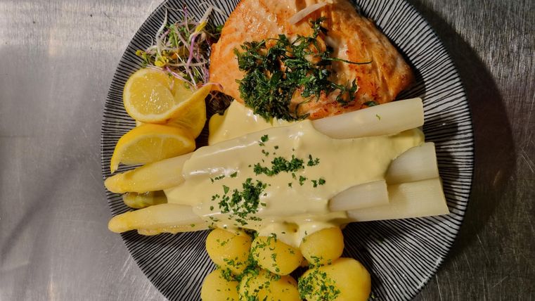 Een bord met gegrilde zalm, asperges en kleine aardappelen. Daarbij zijn schijfjes citroen en verse kruiden gedecoreerd.