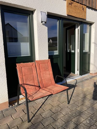 Ein zweisitziger Stuhl in orangefarbenem Stoff steht vor einer Haustür. Die Wände sind weiß und die Fenster haben grüne Rahmen.