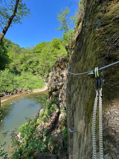 Een mooi wandelpad langs een rivier, omgeven door weelderig groen en hoge rotsen. De lucht is helder en blauw.