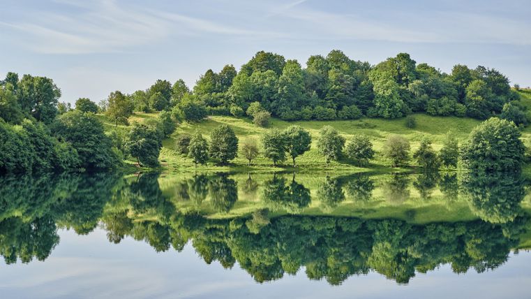 Ein ruhiger See mit spiegelndem Wasser und üppigem Grün drumherum. Die Bäume und Hügel reflektieren sich wunderschön im Wasser.