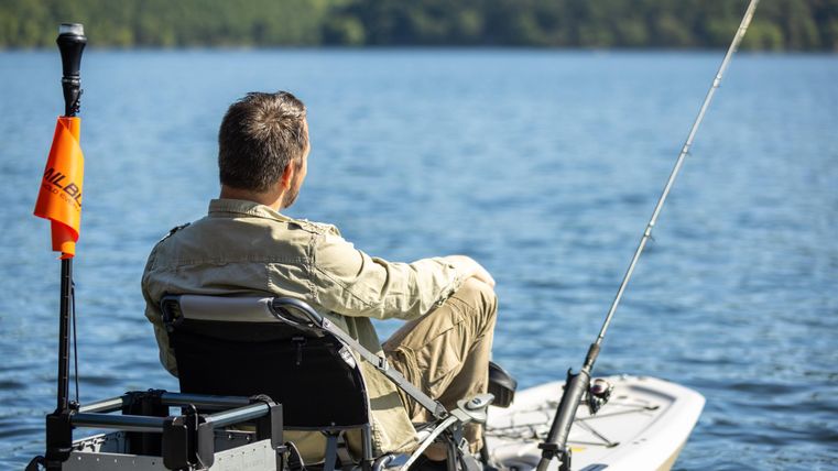 Een man zit op een boot en vist op een rustig water. Het landschap op de achtergrond is groen en pittoresk.
