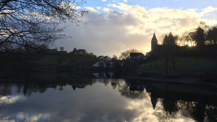 A calm lake with reflective water and a dramatic sky. Trees and a church tower are visible at the shore.