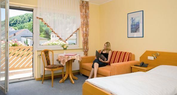 Ein helles Hotelzimmer mit einem orangefarbenen Sofa und einem Tisch. Eine Frau sitzt auf dem Sofa und liest in einem Buch, während das Fenster zum Balkon geöffnet ist.