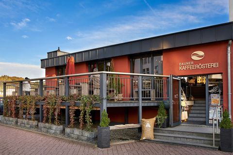 Ein modernes Café mit einer einladenden Terrassenfläche. Die Fassade ist rot, und es gibt einige Pflanzen zur Dekoration.