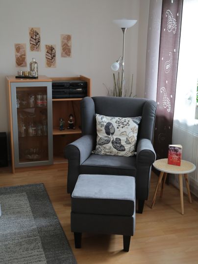 Ein gemütlicher Sessel mit einem Kissen steht neben einem kleinen Beistelltisch. Im Hintergrund sind ein Regal und dekorative Wandbilder zu sehen.