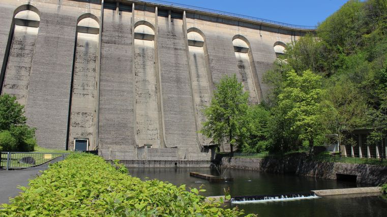 Ein großer Staudamm mit einer steilen, betonten Betonoberfläche. Im Vordergrund befinden sich Bäume und ein ruhiger Wasserlauf.