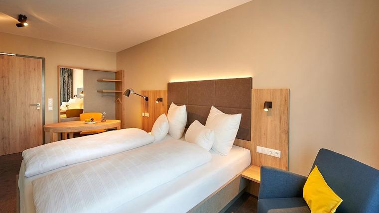 Ein modernes Hotelzimmer mit einem großen Doppelbett und gemütlichen Kissen. Im Hintergrund sind ein Tisch und ein Stuhl sowie ein Spiegel zu sehen.