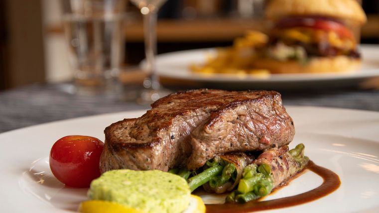 Ein saftiges Steak auf einem Teller, umgeben von grünen Bohnen und einer Soße. Im Hintergrund wird ein Burger mit Pommes serviert.