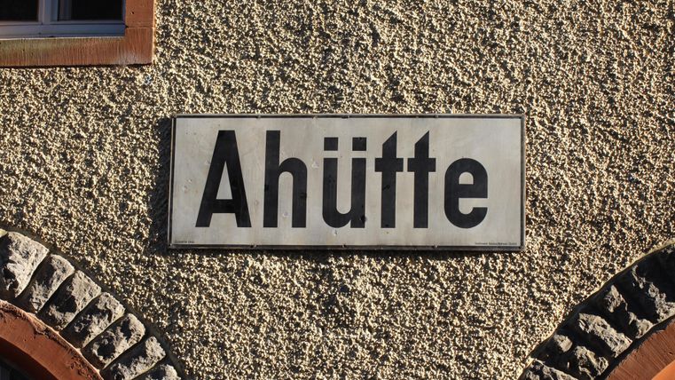 A station sign with the inscription 'Ahütte' hangs on a roughly plastered wall next to a brick arch.