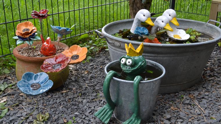 Bunte Keramikblumen und Figuren in Töpfen und Wannen im Garten. Ein Frosch mit Krone sitzt in einem Eimer, Enten schwimmen in einer Wanne.