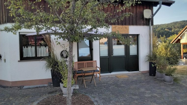 Ein modernes Haus mit einer gepflasterten Einfahrt und einem kleinen Tisch mit Stühlen. Vor dem Haus steht ein Baum und die Umgebung ist grün und freundlich.