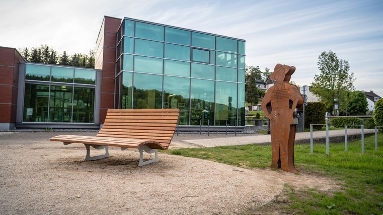 Ein moderner Gebäudekomplex mit großen Glasfenstern. Im Vordergrund steht eine Holzbank und eine Metallfigur, die einen Mann darstellt.