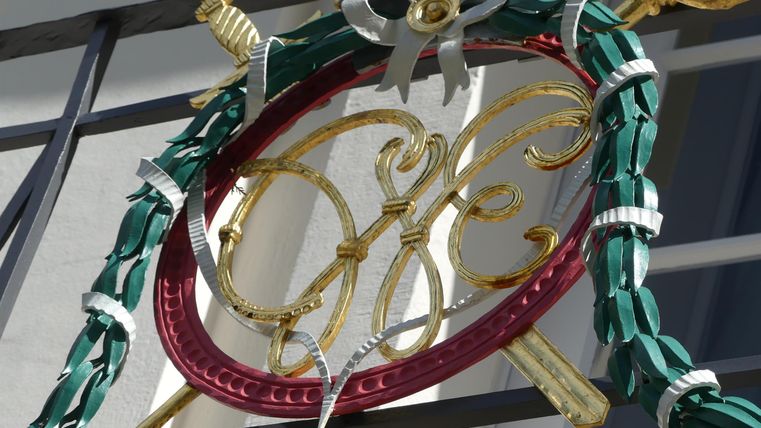 Een decoratief wapen met gouden versieringen en een groene krans. Het hangt aan een zwart hek.