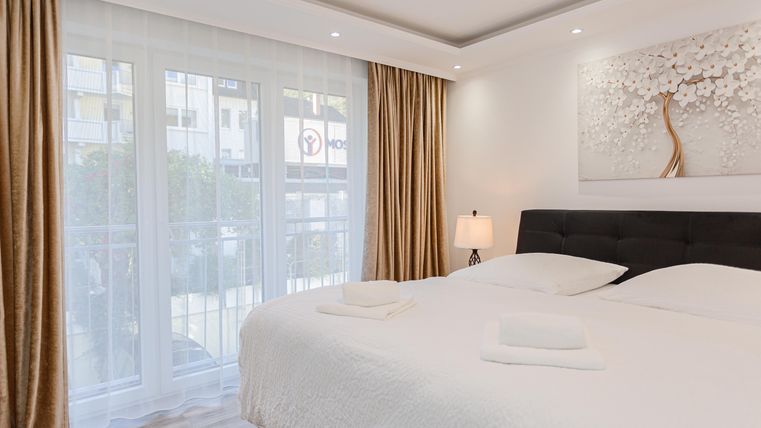 Ein modernes Schlafzimmer mit einem großen Bett und einem Nachttisch. Vorhänge und Fenster sorgen für ein helles Ambiente.