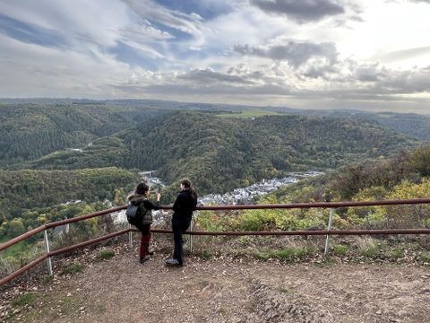 Zwei Personen stehen an einem Aussichtspunkt und blicken auf eine grüne Landschaft. Am Horizont sind Hügel und ein kleiner Ort zu sehen.