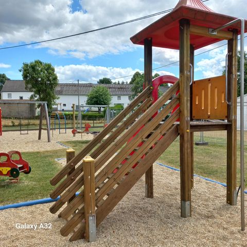 Speelplaats met klimrek en glijbaan, omringd door schommels en wippen. Op de achtergrond zijn bomen en gebouwen te zien. De lucht is deels bewolkt.
