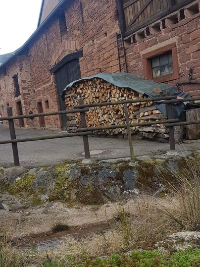 Ein altes, rotes Gebäude mit Holzstapel im Vordergrund. Die Umgebung ist ländlich und wirkt ruhig.