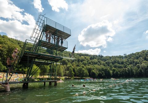 Ein Sprungturm steht neben einem klaren Wassersee, umgeben von Bäumen. Mehrere Schwimmer genießen das erfrischende Wasser.