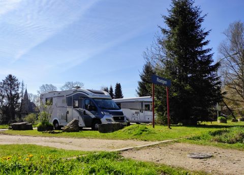 Ein schöner Campingplatz mit zwei Wohnmobilen und einer grünen Umgebung. Im Hintergrund sind Bäume und ein Weg sichtbar.