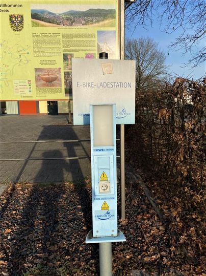 Eine E-Bike-Ladestation steht an einem Weg. Im Hintergrund sind Informationen und Karten zu sehen.